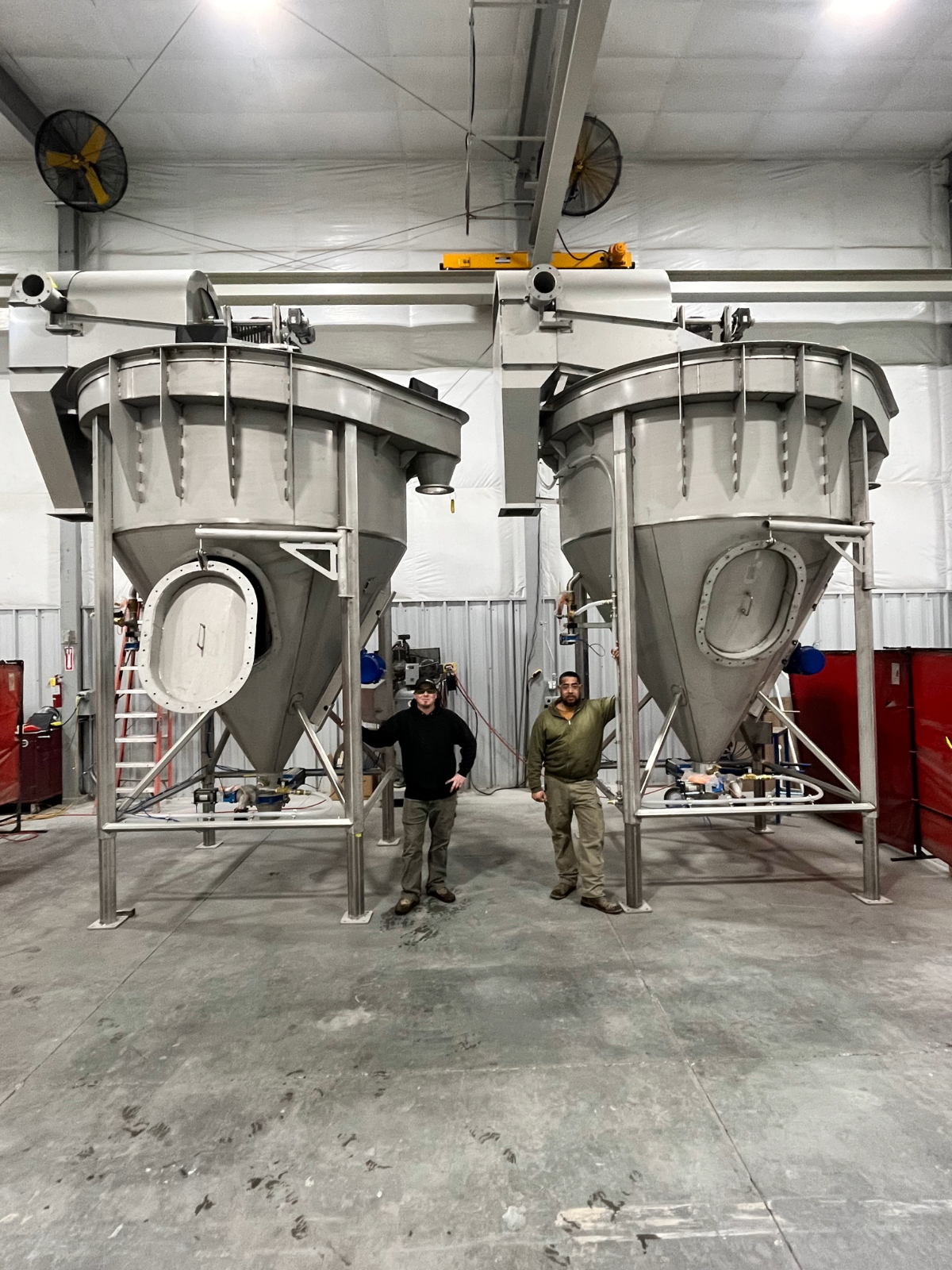Two Water-Re fabricators standing between two stainless steel solids extraction systems under construction in the Burley, Idaho shop